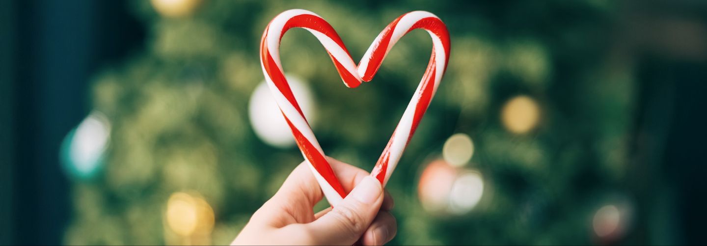 Hand hält zwei rot-weiß gestreifte Zuckerstangen, die zusammen ein Herz formen, unscharfer Hintergrund mit Lichtern.