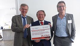 Drei Personen stehen zusammen und halten ein großes Schild mit der Aufschrift 'Wir helfen Paul' und dem Betrag '26.776 €'. Die Personen tragen Anzüge.