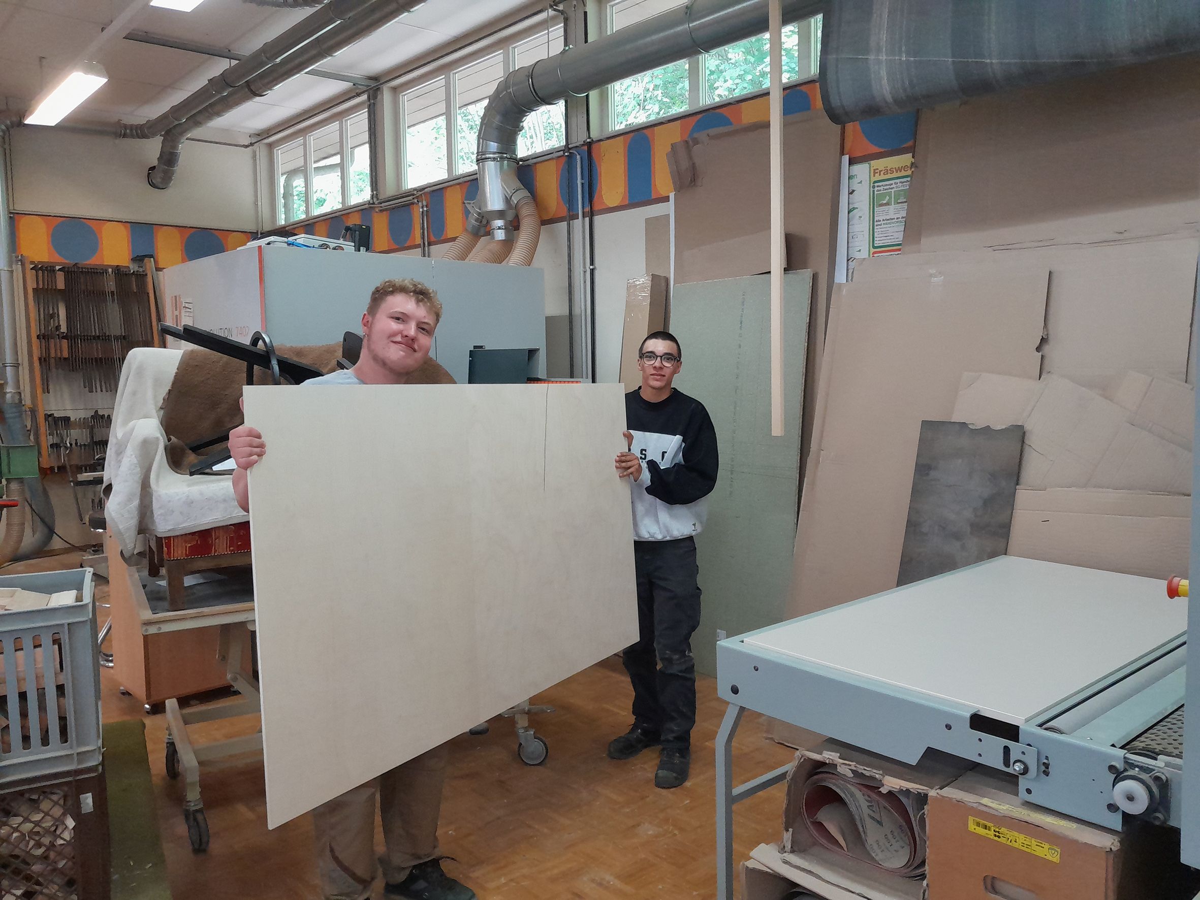 Zwei junge Männer mit einer Holzplatte stehen in einer Schreinerei