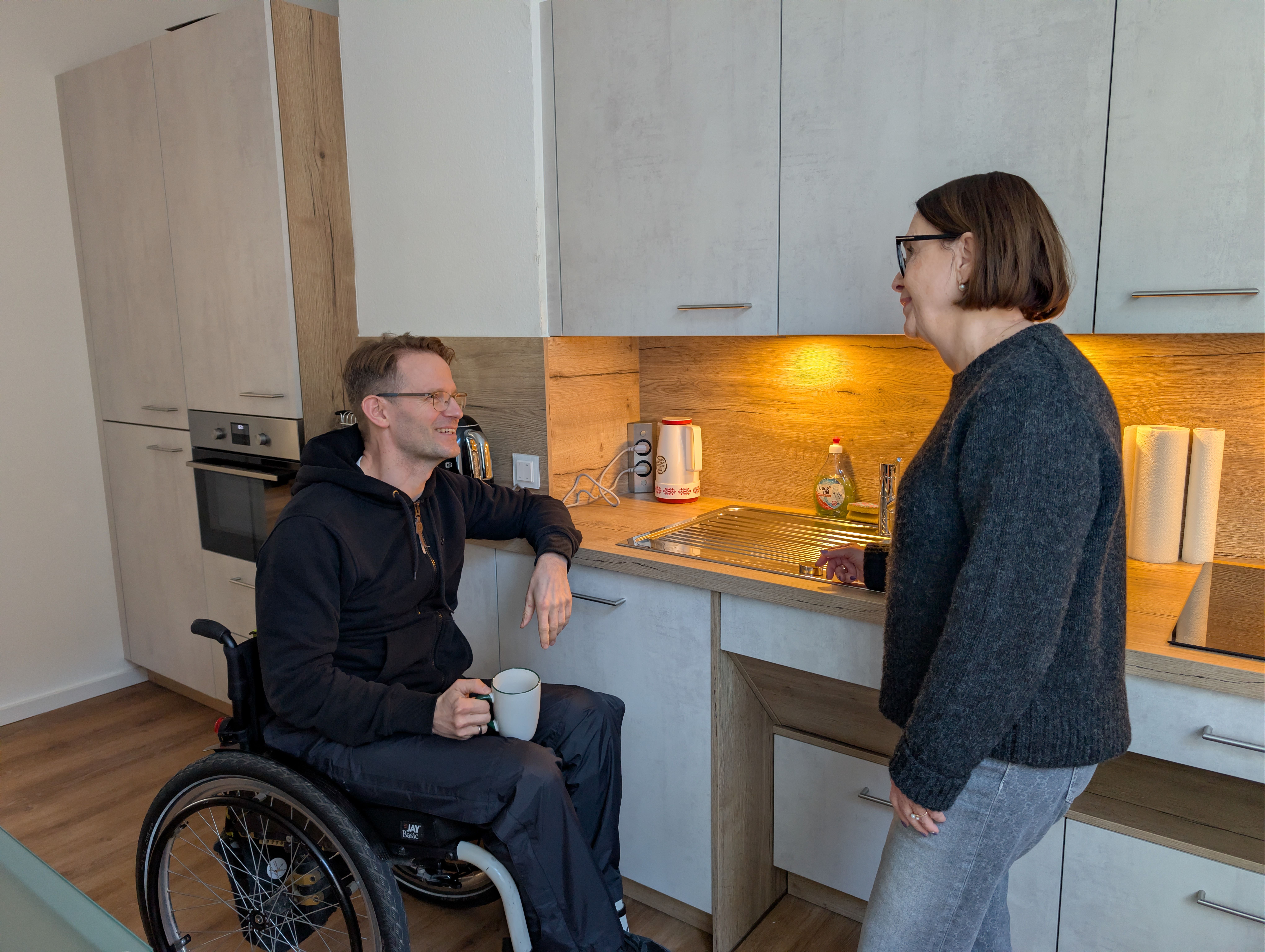 Ein Mann im Rollstuhl und eine Frau unterhalten sich vor einer Küchenzeile.