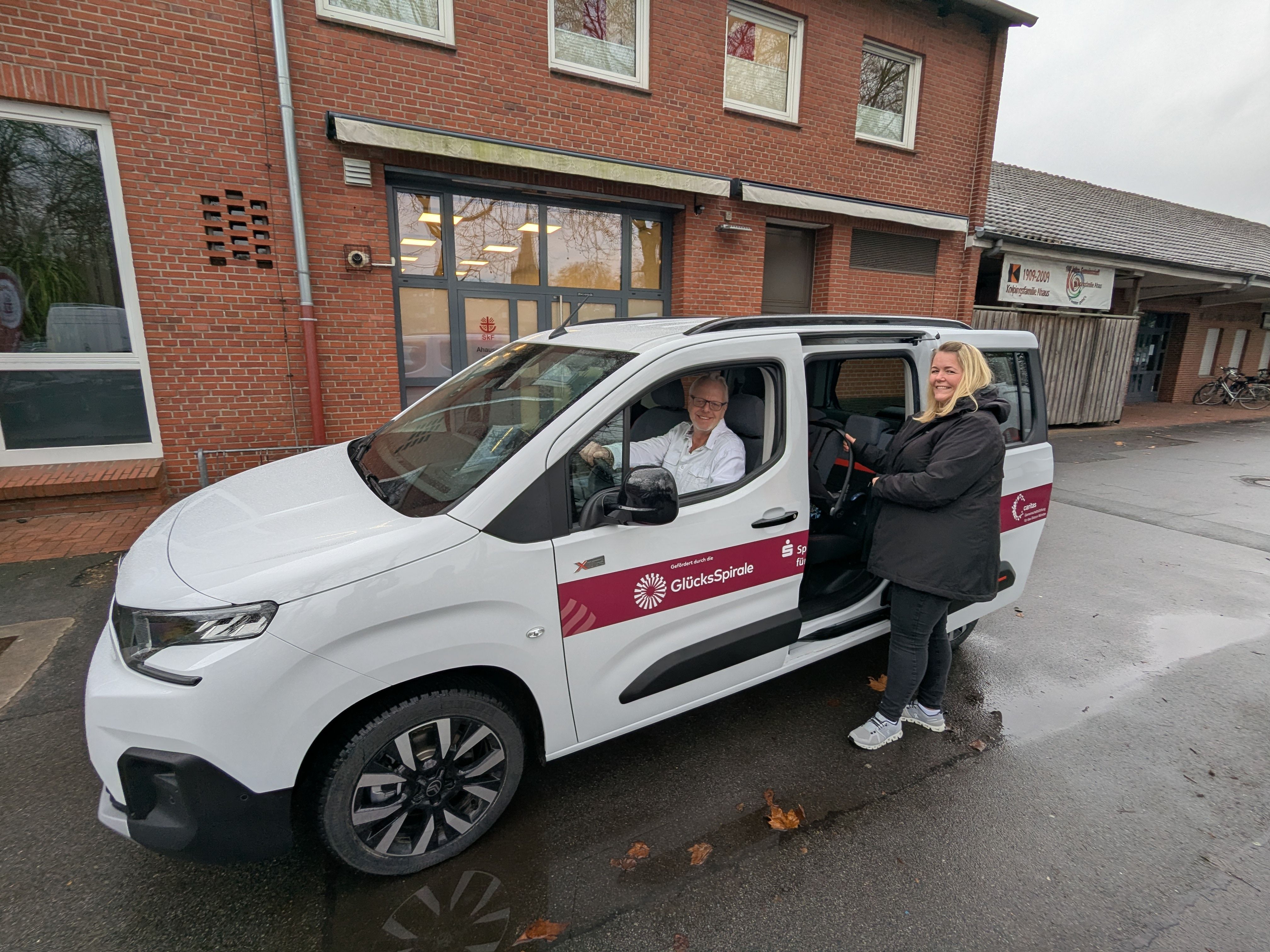Ein Mann fährt einen Kleintransporter, eine Frau hält bei offener Fahrzeugtür einen Maxicosi in der Hand