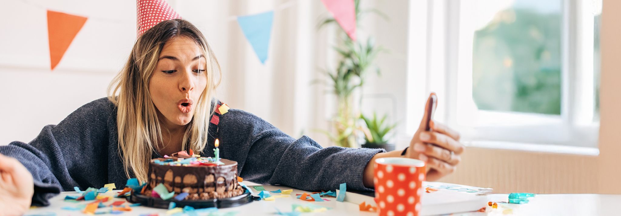 Person mit Partyhut sitzt an Tisch mit Schokoladenkuchen und bunter Dekoration und hält Smartphone zum Selfie in der Hand.