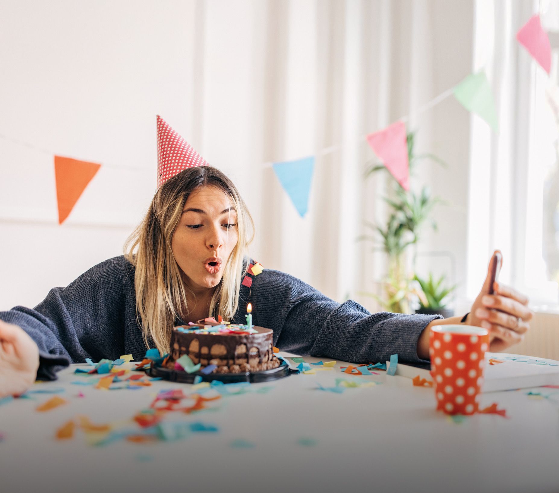Person mit Partyhut sitzt an Tisch mit Schokoladenkuchen und bunter Dekoration und hält Smartphone zum Selfie in der Hand.